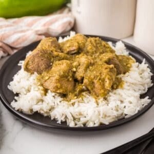 A black plate with white rice topped with chunks of pork chili verde in a green sauce, placed on a white surface with a striped cloth and jars in the background.