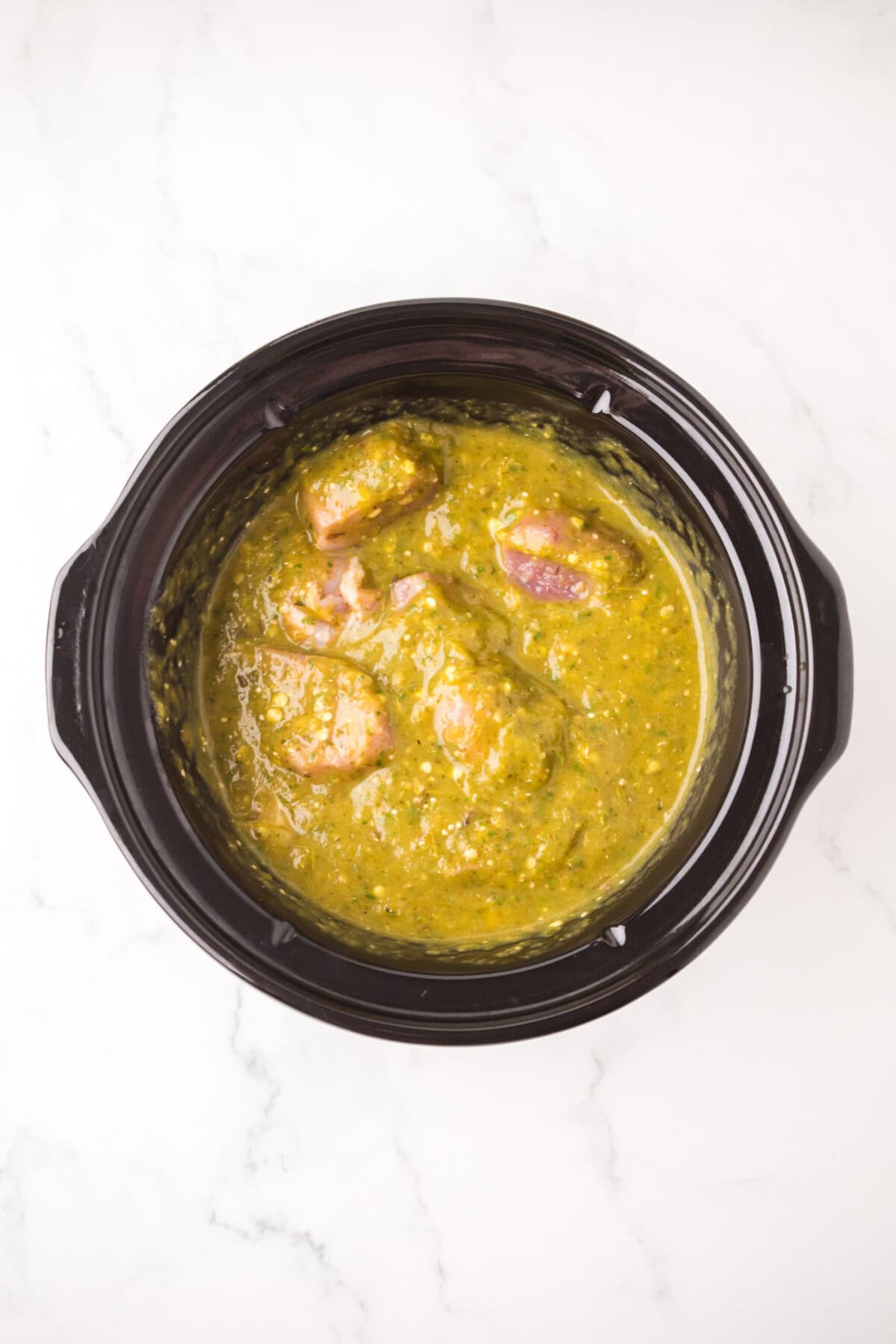A black slow cooker filled with pieces of raw pork chili verde, coated in a vibrant green sauce, sits on a white marble surface.