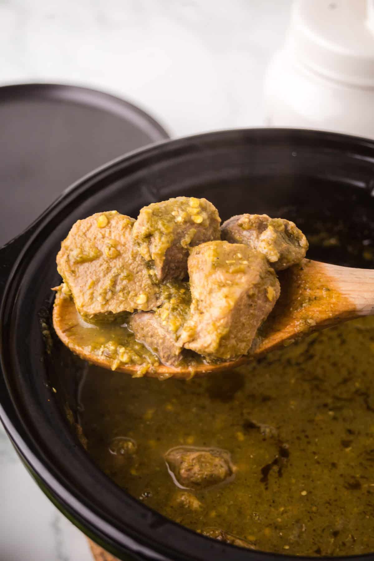 A wooden spoon lifts chunks of cooked pork in a greenish-brown pork chili verde sauce from a slow cooker, with the lid resting nearby.