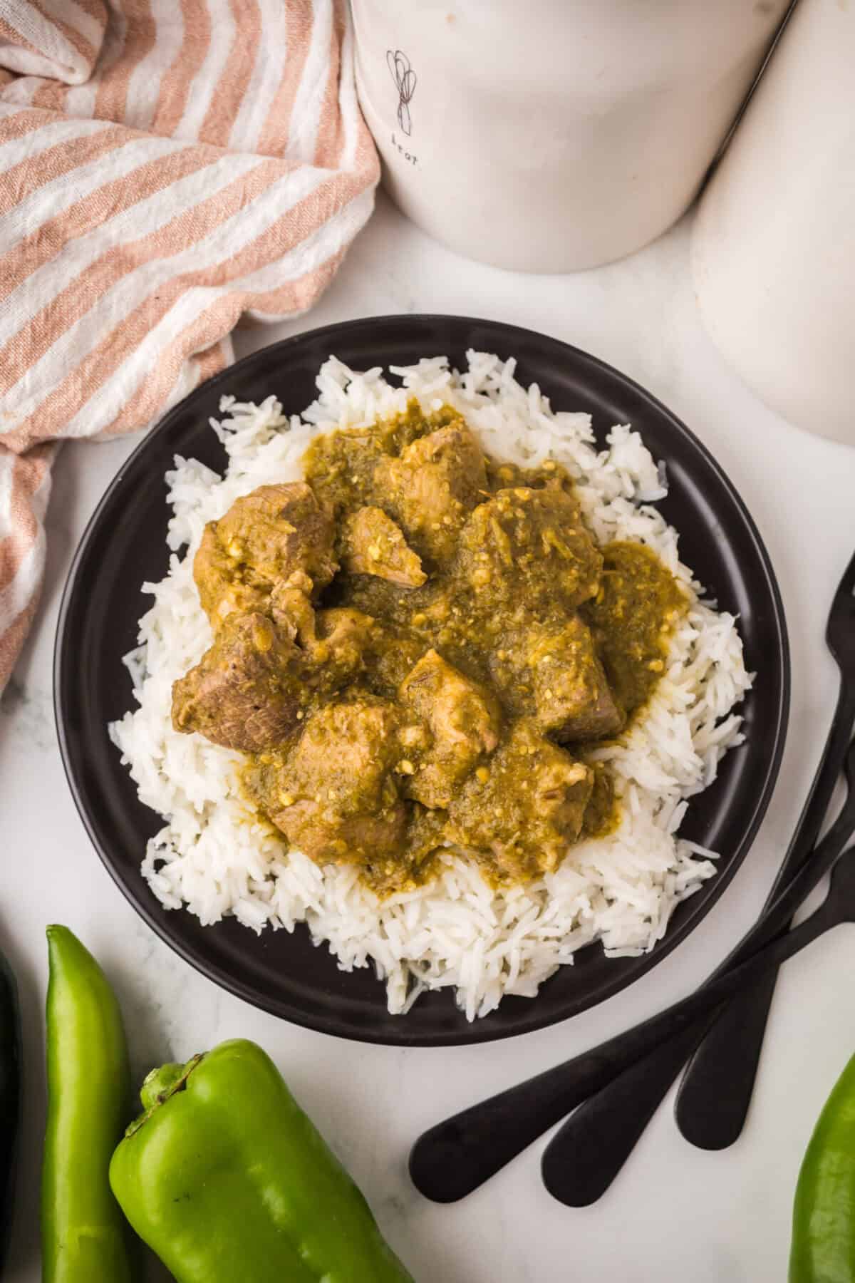 A black plate filled with white rice topped with pieces of pork chili verde in a greenish curry sauce, placed on a white surface with green peppers, utensils, and a striped cloth nearby.