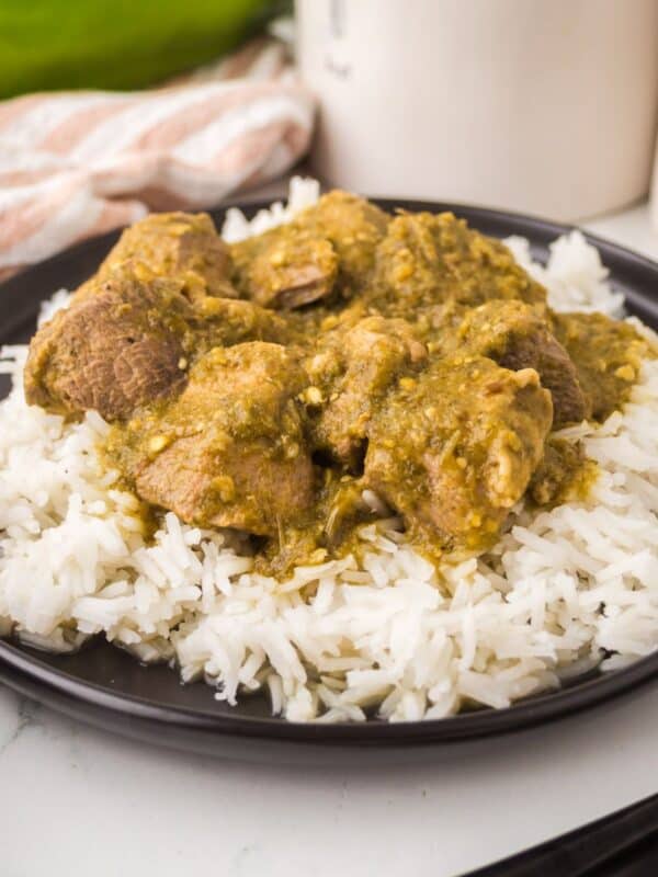 A black plate with white rice topped with chunks of pork chili verde in a green sauce, placed on a white surface with a striped cloth and jars in the background.