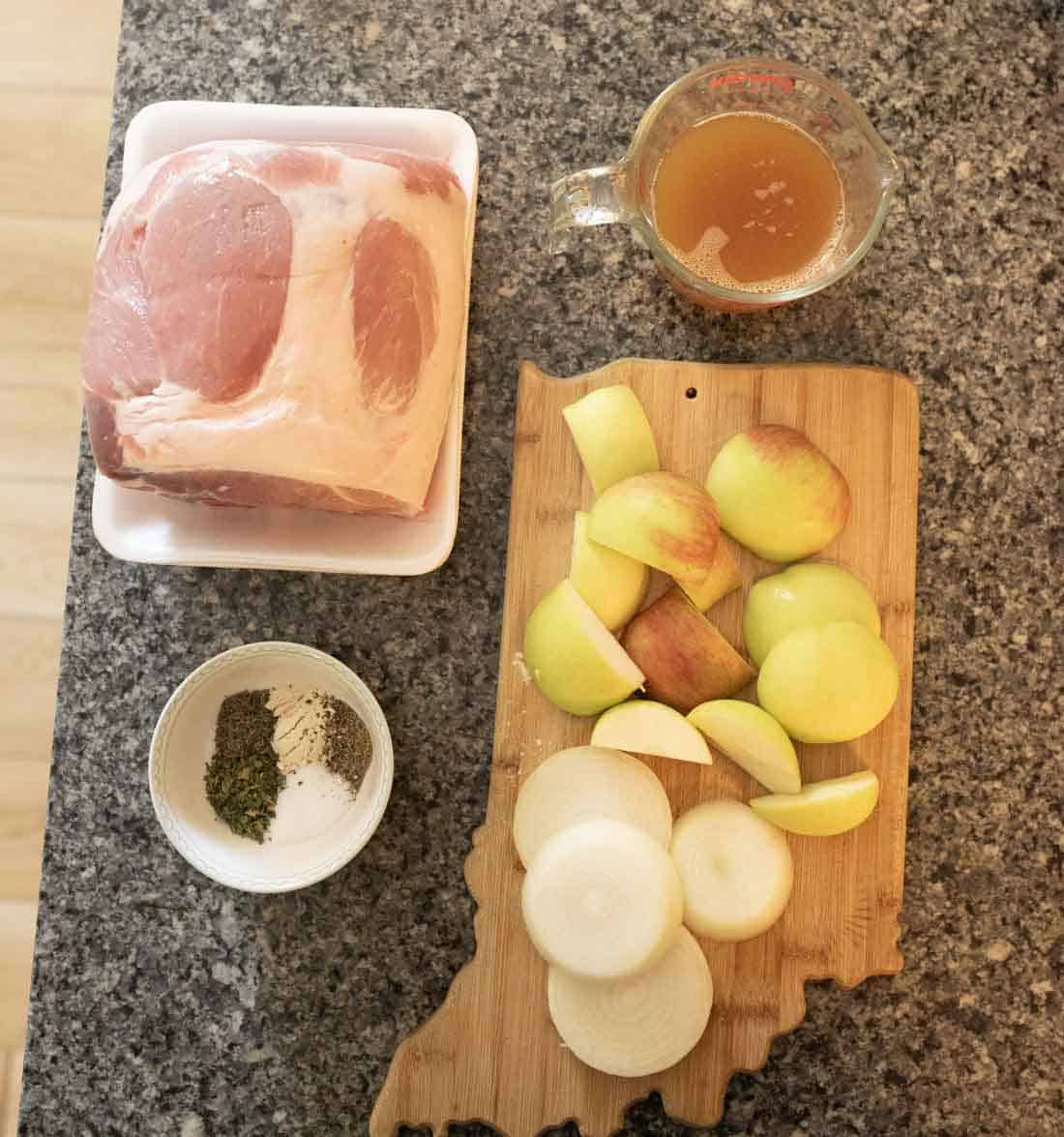 A raw pork roast, a bowl of herbs and pepper, a measuring cup of broth, and a wooden board with sliced apples and onions are arranged on a kitchen counter.