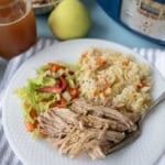 A white plate with shredded pork, vegetable rice, and a lettuce salad. A fork is on the plate. In the background are a glass of iced tea, a pear, a bowl of shredded pork, and a pressure cooker.