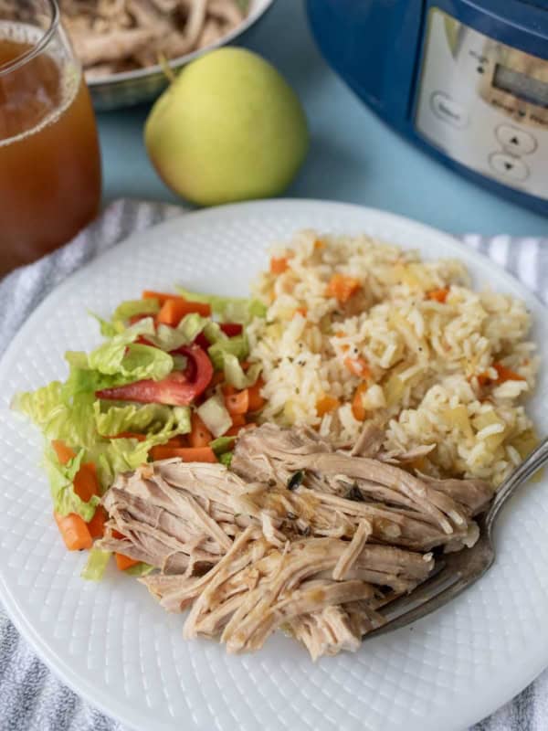 A white plate with shredded pork, vegetable rice, and a lettuce salad. A fork is on the plate. In the background are a glass of iced tea, a pear, a bowl of shredded pork, and a pressure cooker.