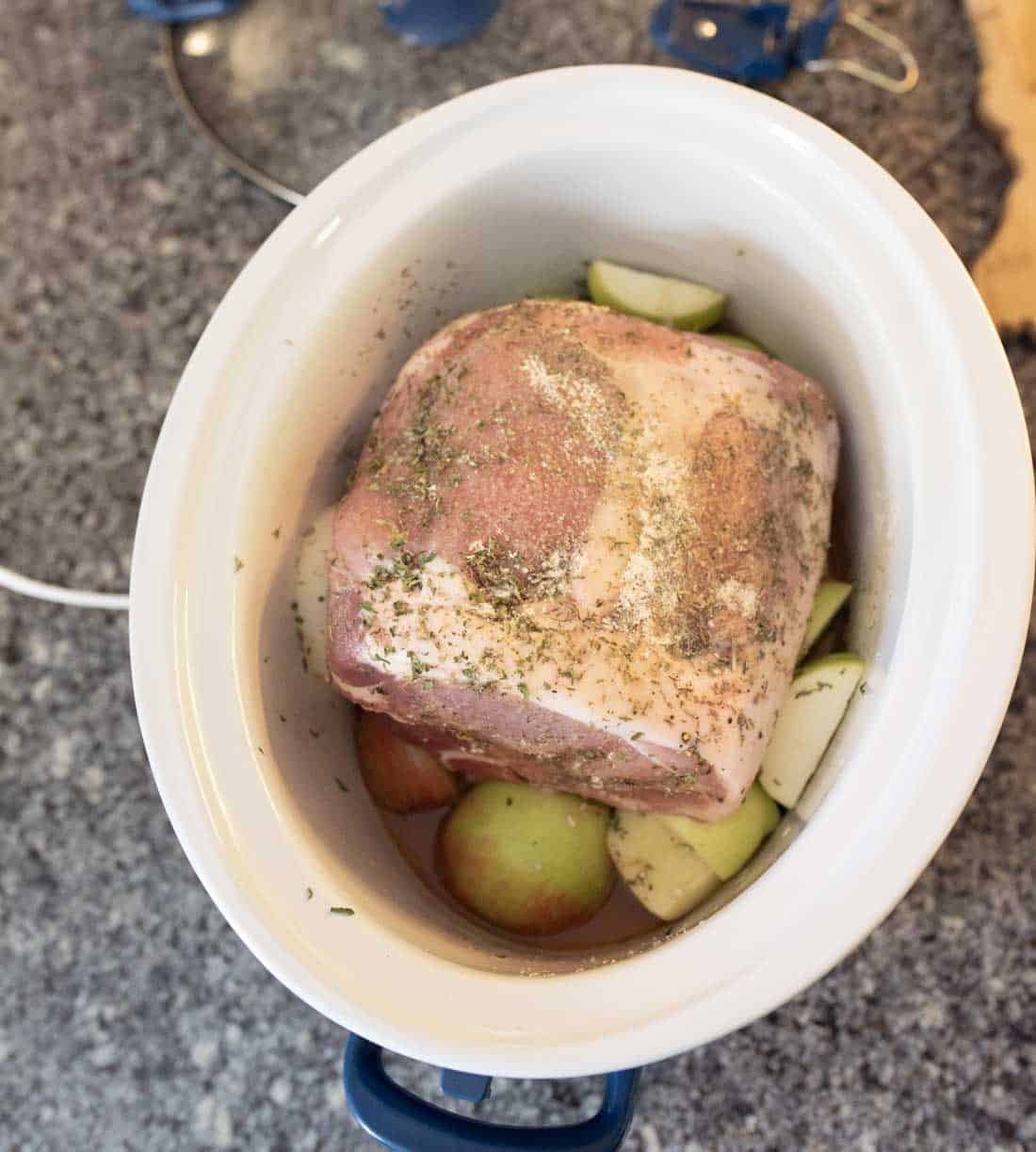 A raw seasoned pork roast sits in a white slow cooker on a grey countertop, surrounded by sliced green apples and liquid at the bottom.