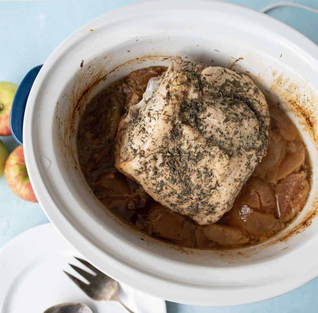A cooked pork roast seasoned with herbs sits on top of apples and onions inside a white slow cooker, with a plate and serving utensils nearby.