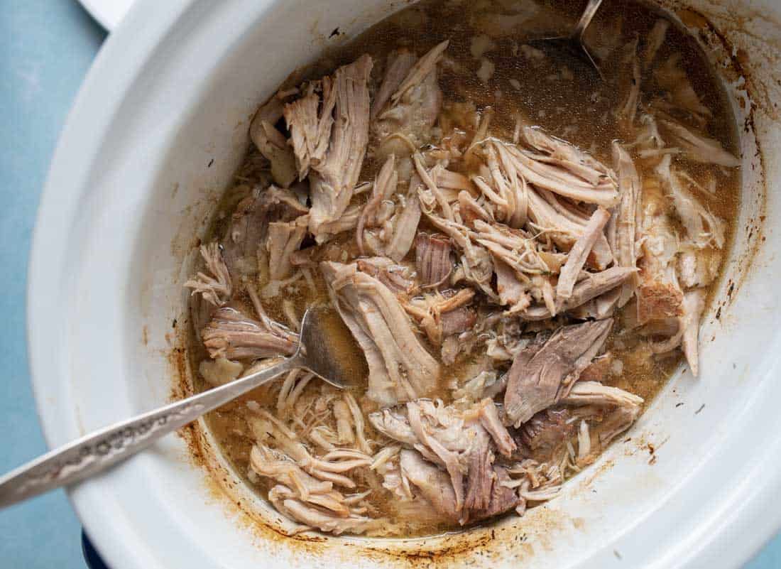 Shredded cooked pork in a slow cooker with two metal forks resting in the juicy, tender meat. The meat appears moist and flavorful, with visible broth and pulled pieces.