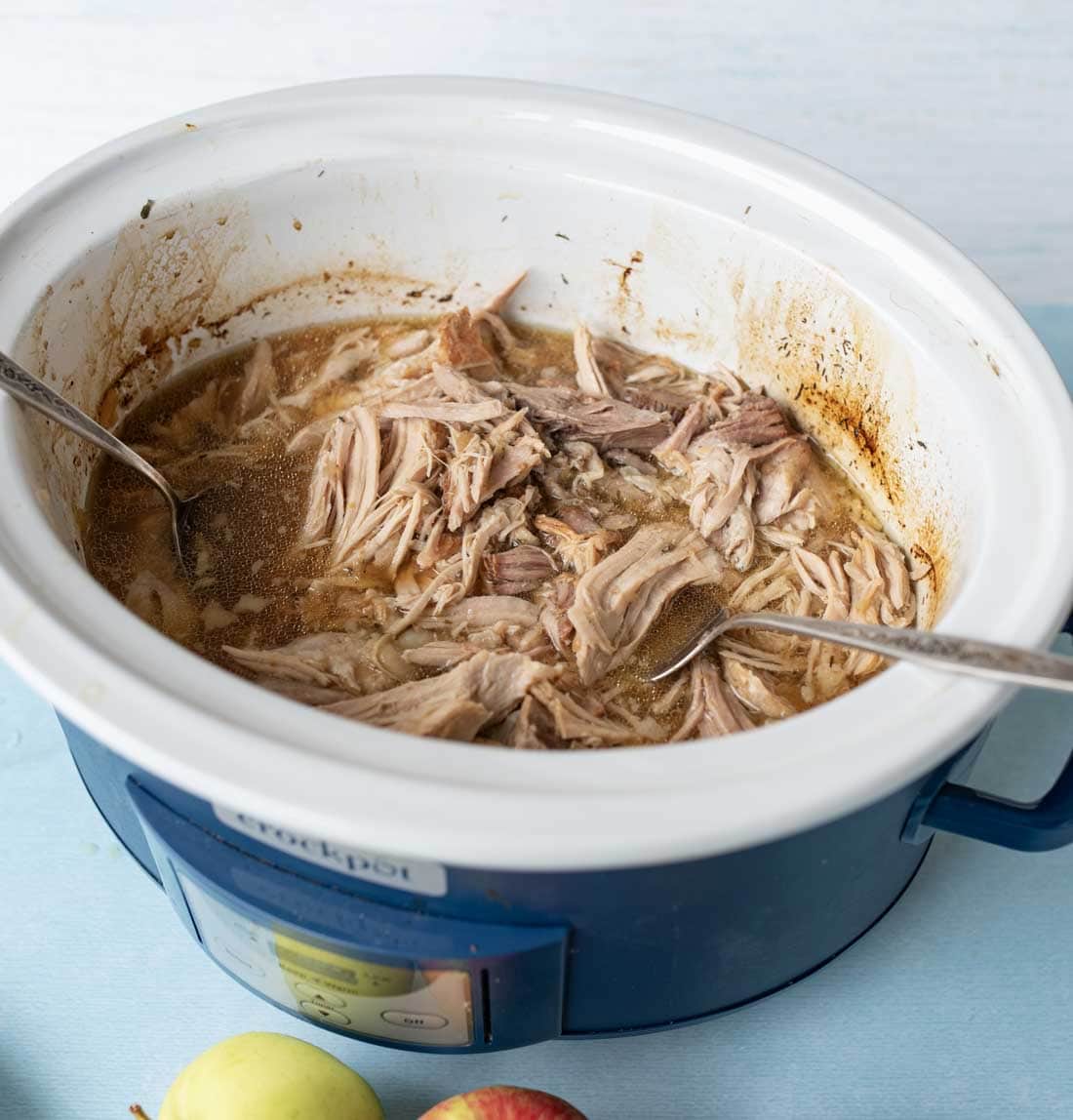 Shredded pork in a blue slow cooker with two forks, surrounded by broth. The slow cooker sits on a light blue surface with apples placed nearby.