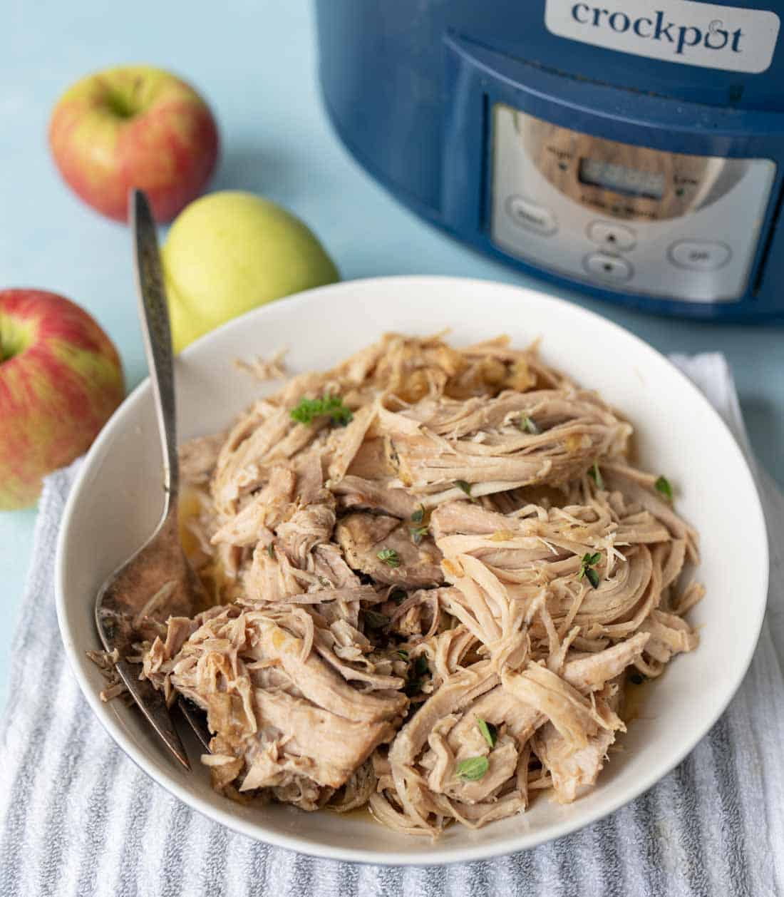 A bowl of shredded pork garnished with herbs sits on a striped towel, next to a spoon. Two apples and a blue crockpot are in the background.