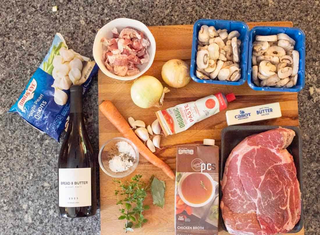 A selection of beef stew ingredients on a counter, including a large cut of beef, two onions, carrot, garlic, mushrooms, bacon, tomato paste, beef broth, butter, herbs, salt, pepper, and a bottle of red wine.