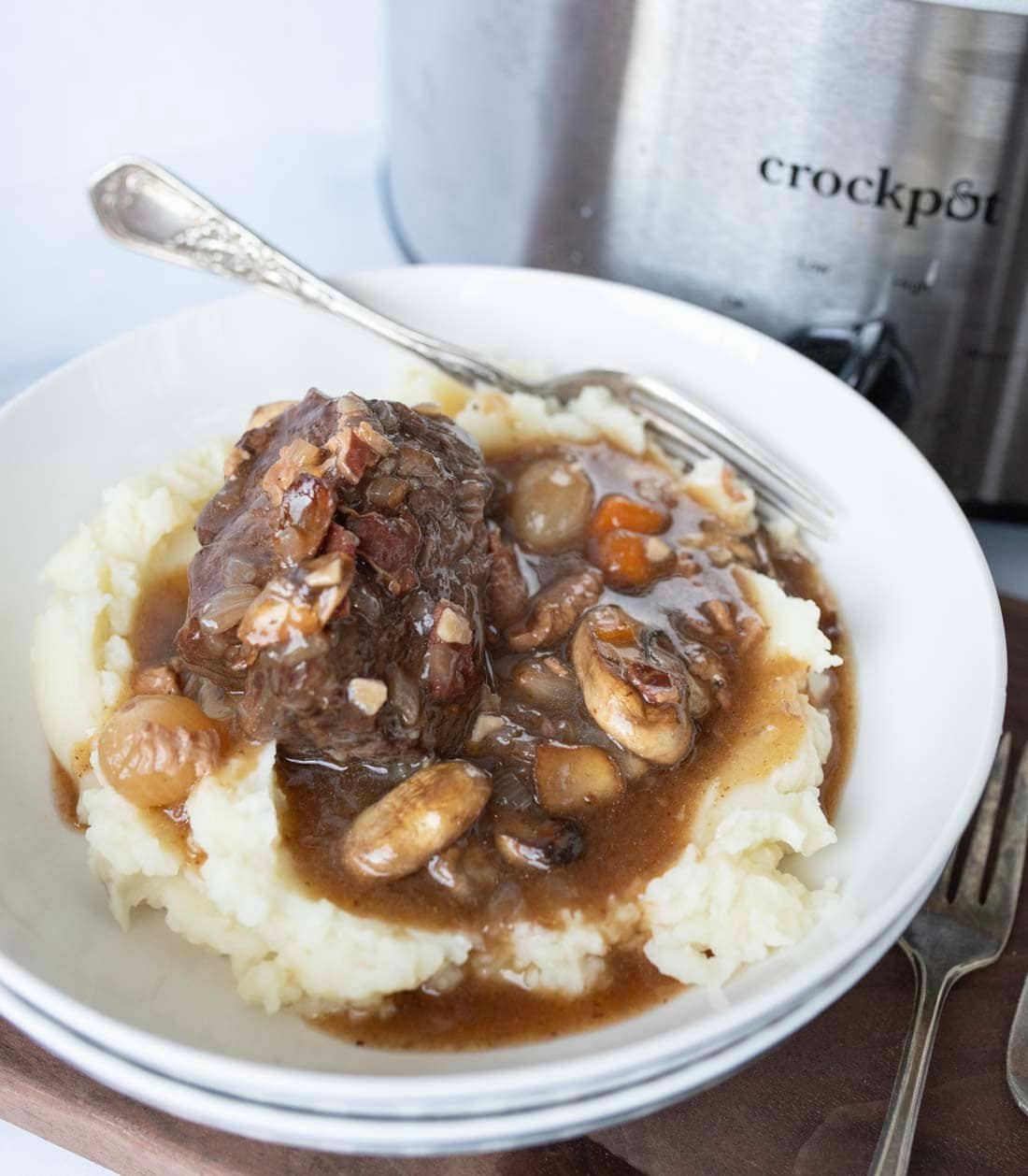 A white bowl filled with mashed potatoes topped with beef stew containing onions, mushrooms, and carrots, with a fork and a crockpot in the background.