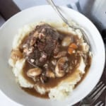 A bowl of mashed potatoes topped with slow cooker beef bourguignon containing mushrooms, carrots, onions, and a rich brown gravy. A silver fork rests on the edge of the bowl.