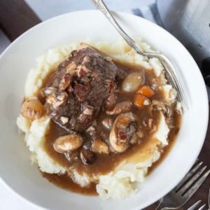 A bowl of mashed potatoes topped with slow cooker beef bourguignon containing mushrooms, carrots, onions, and a rich brown gravy. A silver fork rests on the edge of the bowl.