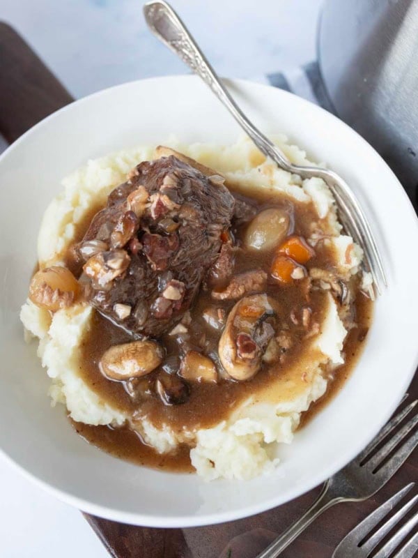 A bowl of mashed potatoes topped with slow cooker beef bourguignon containing mushrooms, carrots, onions, and a rich brown gravy. A silver fork rests on the edge of the bowl.