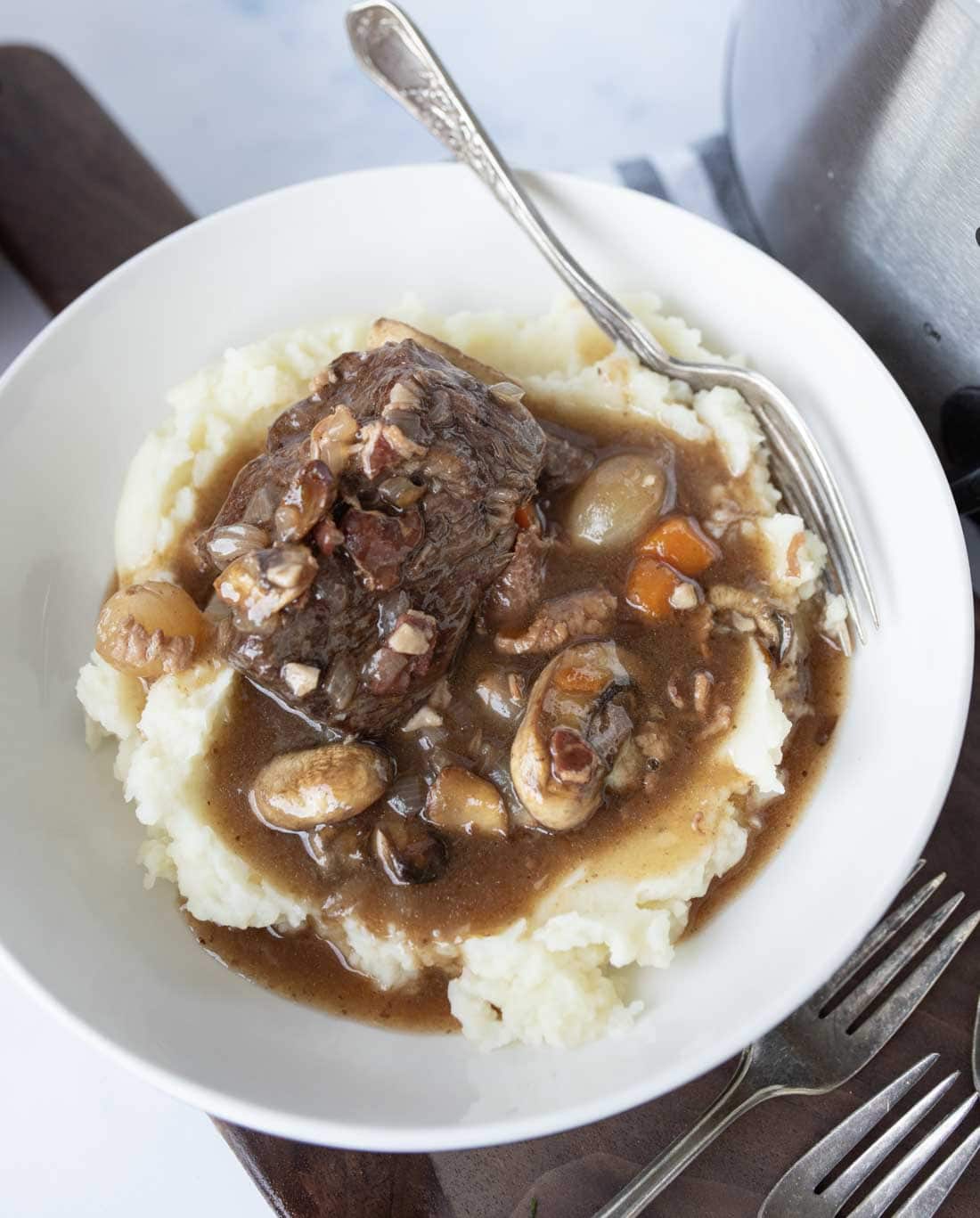 A bowl of mashed potatoes topped with slow cooker beef bourguignon containing mushrooms, carrots, onions, and a rich brown gravy. A silver fork rests on the edge of the bowl.