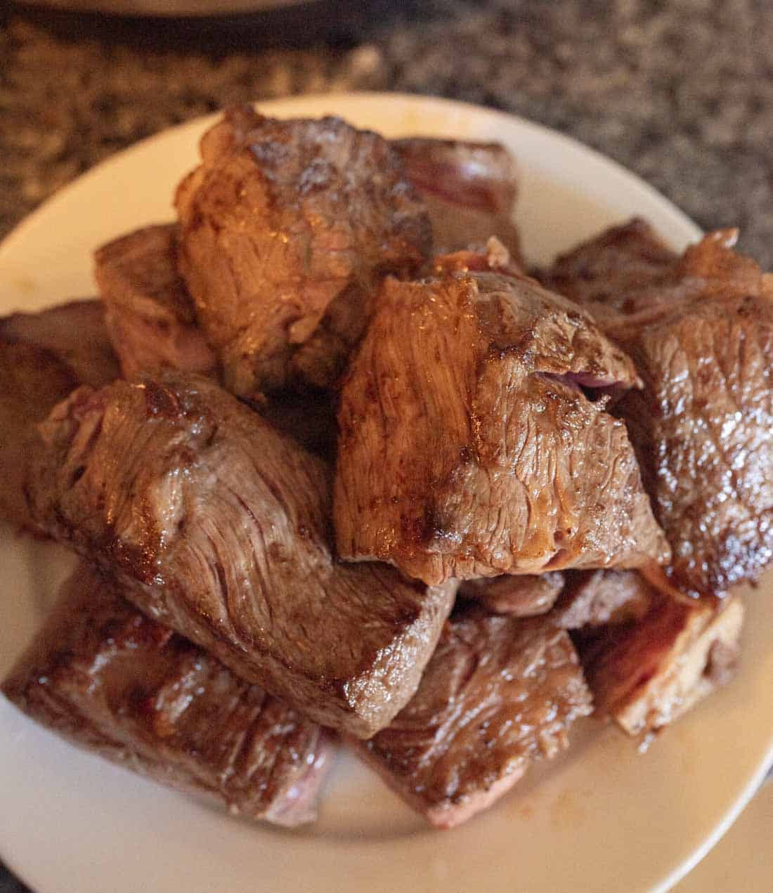 A white plate piled with several pieces of cooked, browned steak, placed on a granite countertop. The meat appears juicy and tender with a seared exterior.