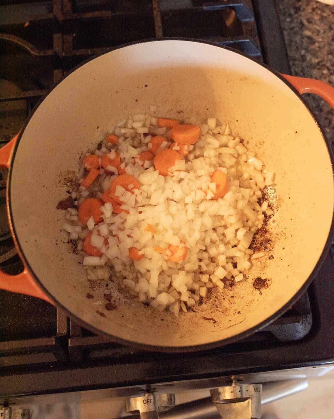 A Dutch oven on a stovetop with diced onions and sliced carrots sautéing, showing browned bits on the bottom of the pot.