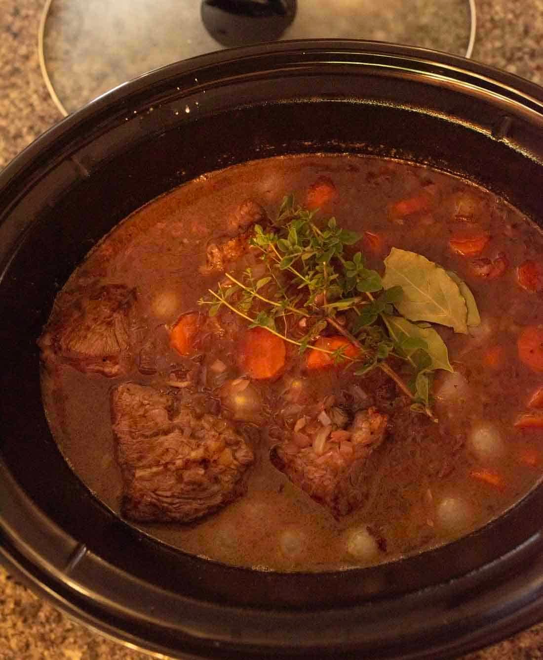 A slow cooker filled with beef stew, featuring chunks of beef, sliced carrots, pearl onions, broth, and garnished with fresh thyme and bay leaves.