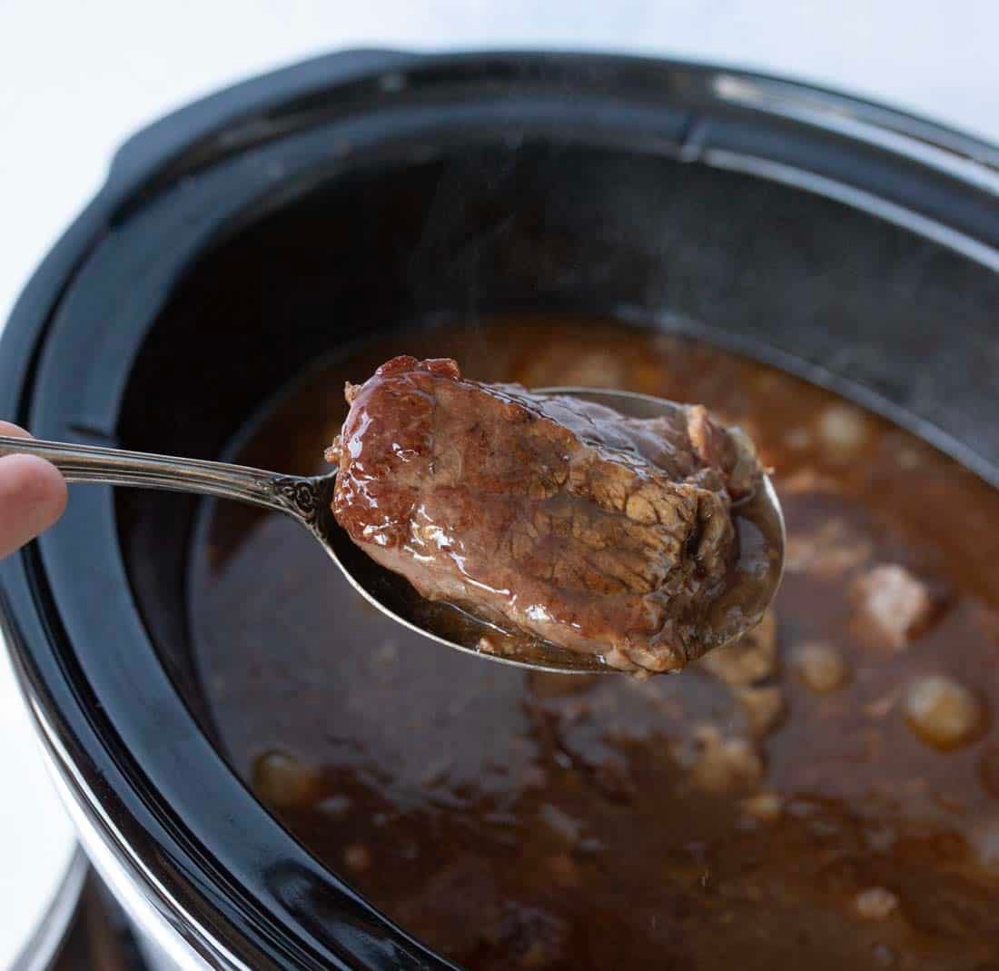 A spoon holding a piece of cooked beef over a slow cooker filled with rich, brown broth and chunks of meat. Steam is rising from the food, indicating it is hot and freshly cooked.