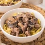 A white bowl filled with slow cooker beef stroganoff—egg noodles topped with tender beef and sliced mushrooms in a creamy sauce—sits on a woven placemat. Another bowl of the same dish is partially visible in the background.