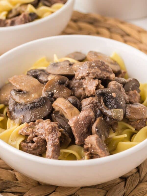 A white bowl filled with slow cooker beef stroganoff—egg noodles topped with tender beef and sliced mushrooms in a creamy sauce—sits on a woven placemat. Another bowl of the same dish is partially visible in the background.