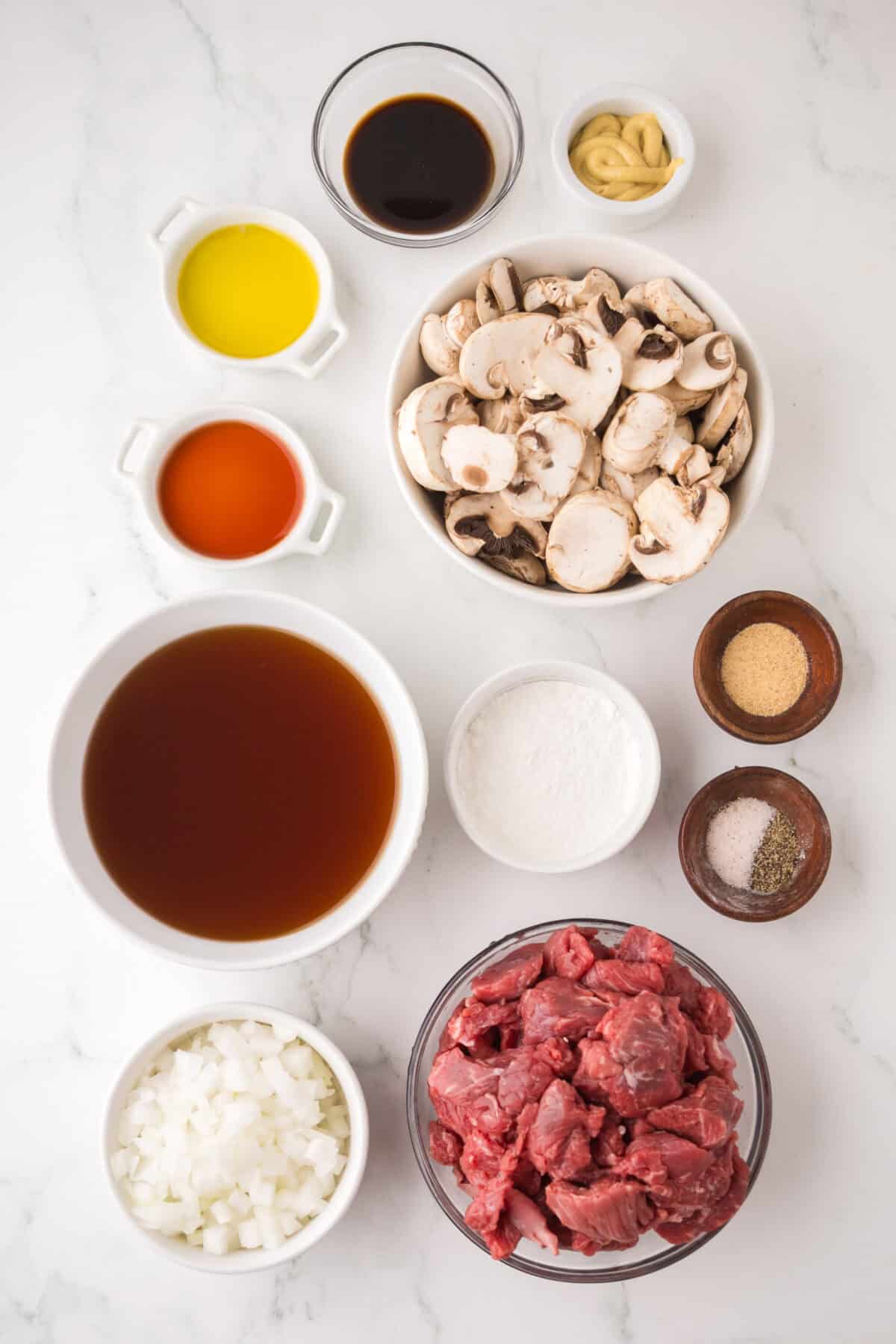 Top-down view of ingredients for slow cooker beef stroganoff on a white surface: sliced mushrooms, cubed beef, diced onions, beef broth, olive oil, Worcestershire sauce, Dijon mustard, flour, garlic powder, salt, pepper, and red vinegar in separate bowls.