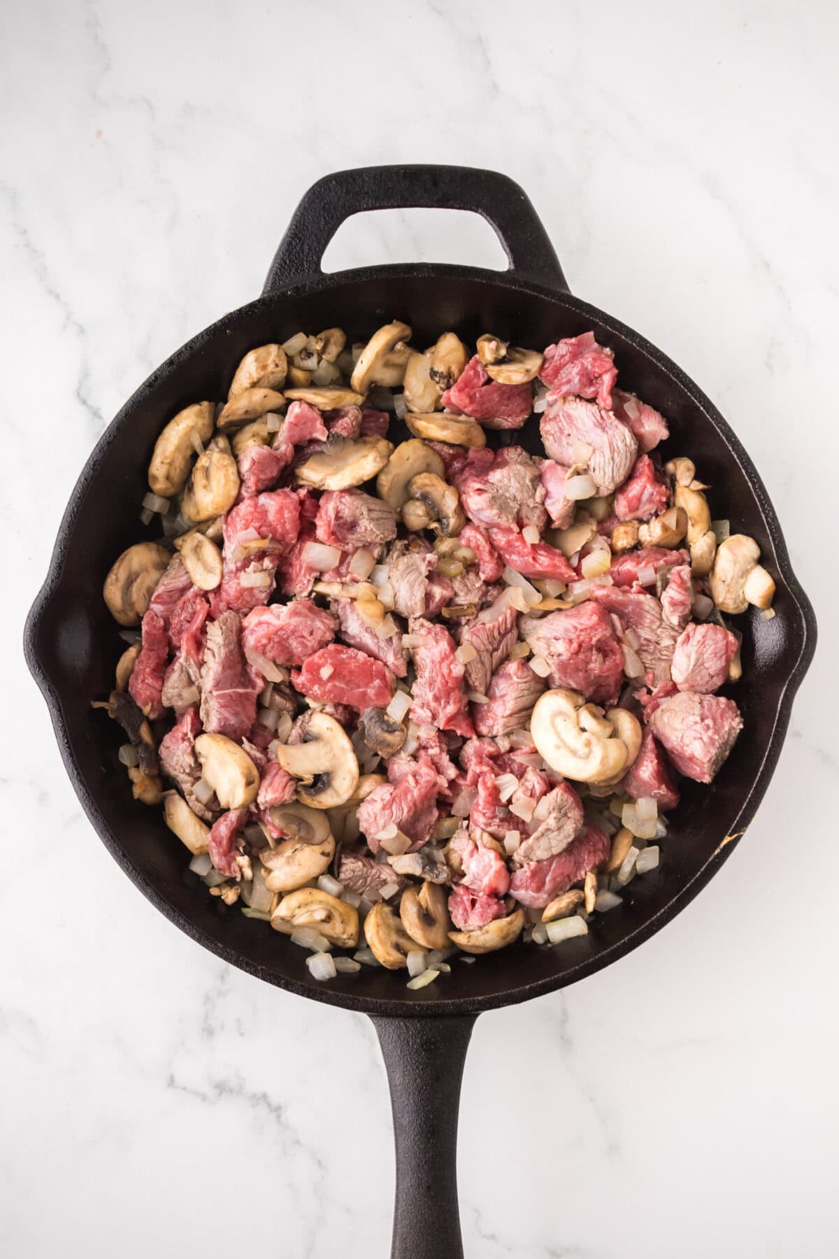 A cast iron skillet filled with sliced beef, mushrooms, and chopped onions being sautéed on a white marble surface—perfect ingredients for a classic slow cooker beef stroganoff.