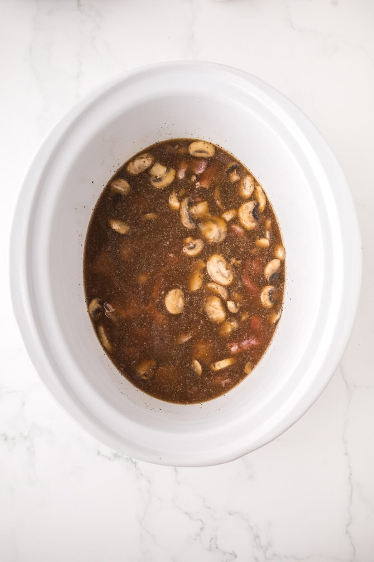 A white slow cooker filled with a mixture of sliced mushrooms and brown broth for slow cooker beef stroganoff, sitting on a white marble surface.
