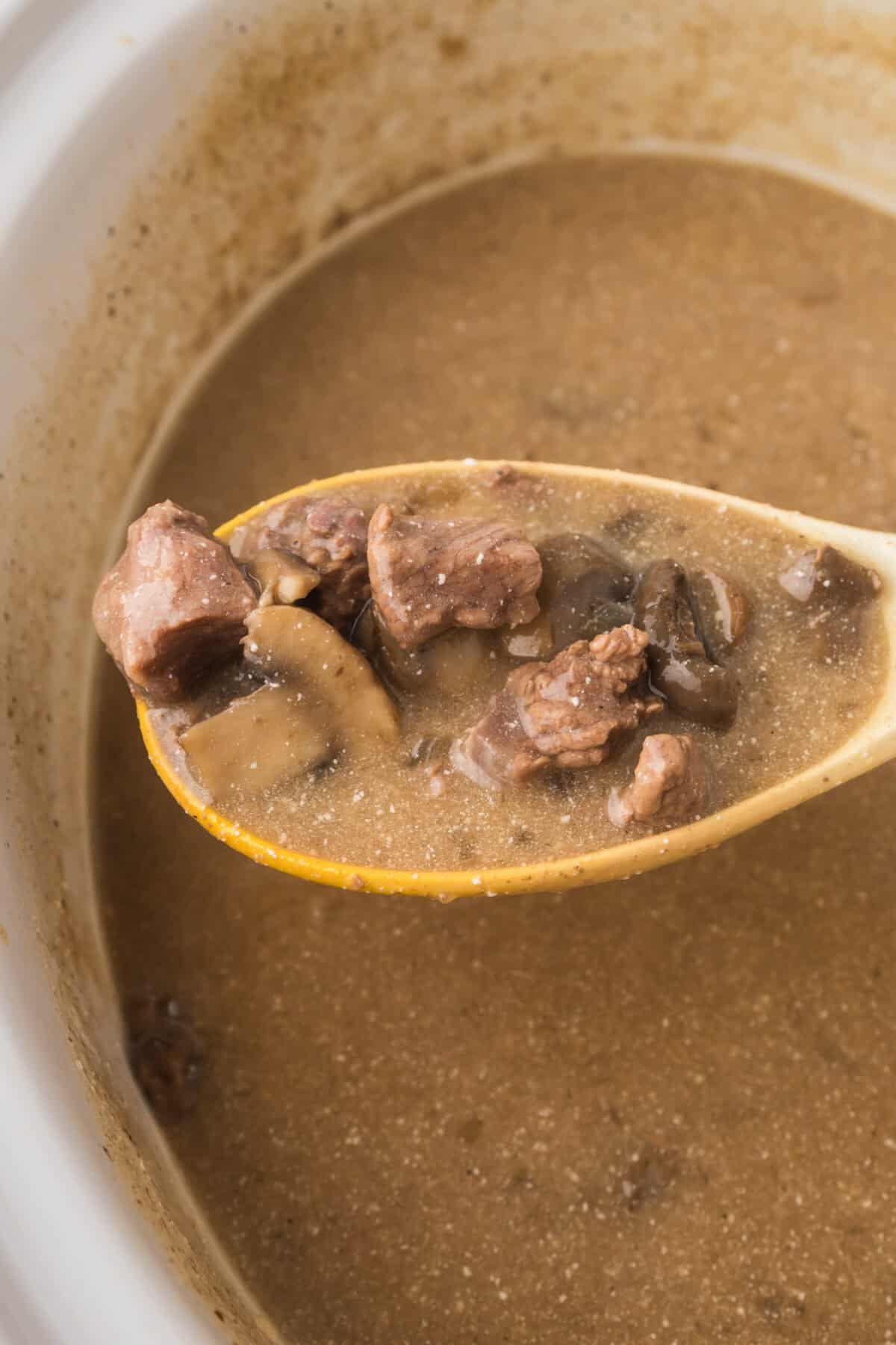 A close-up of a wooden spoon holding pieces of slow cooker beef stroganoff with mushrooms in a brown broth, above a pot of hearty soup or stew.
