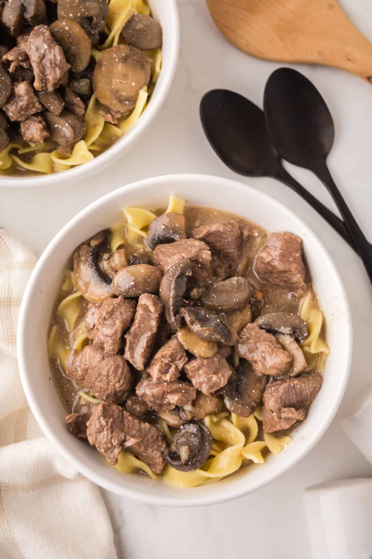 A bowl of slow cooker beef stroganoff features tender chunks of beef, sliced mushrooms, and egg noodles. Another partial bowl is visible, with two black spoons and a wooden spatula nearby on a white surface.