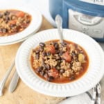 A bowl of chili with ground meat, black beans, and tomatoes sits on a white plate with a spoon. Another bowl and a crockpot are visible in the background on a wooden surface.