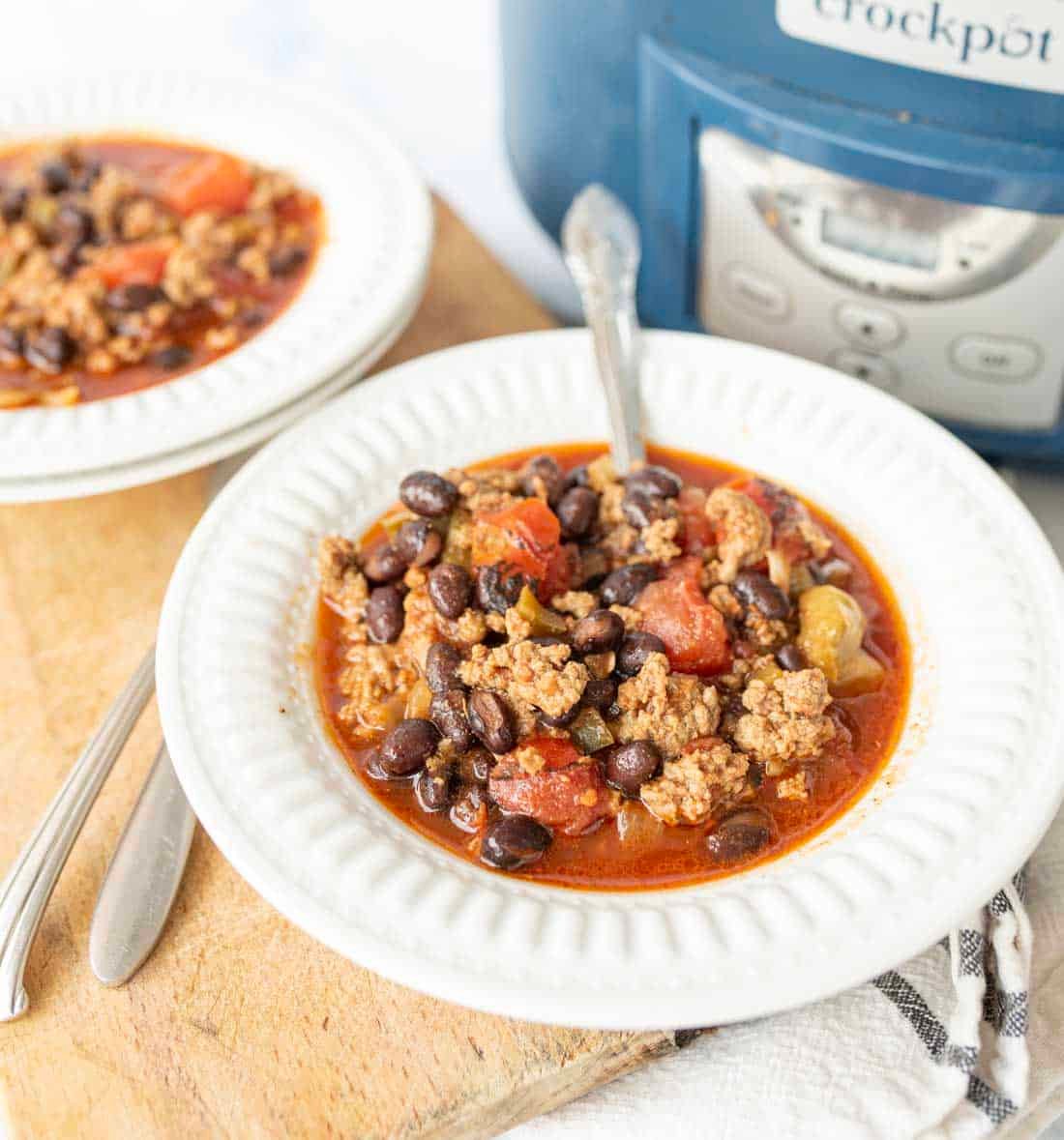 A bowl of chili with ground meat, black beans, and tomatoes sits on a white plate with a spoon. Another bowl and a crockpot are visible in the background on a wooden surface.