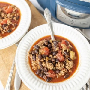 A bowl of black bean chili with ground meat, black beans, and tomatoes sits on a white plate next to a slow cooker labeled crockpot. A spoon is in the bowl and another bowl of chili is nearby.