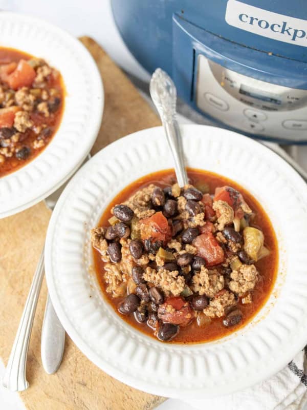 A bowl of black bean chili with ground meat, black beans, and tomatoes sits on a white plate next to a slow cooker labeled crockpot. A spoon is in the bowl and another bowl of chili is nearby.