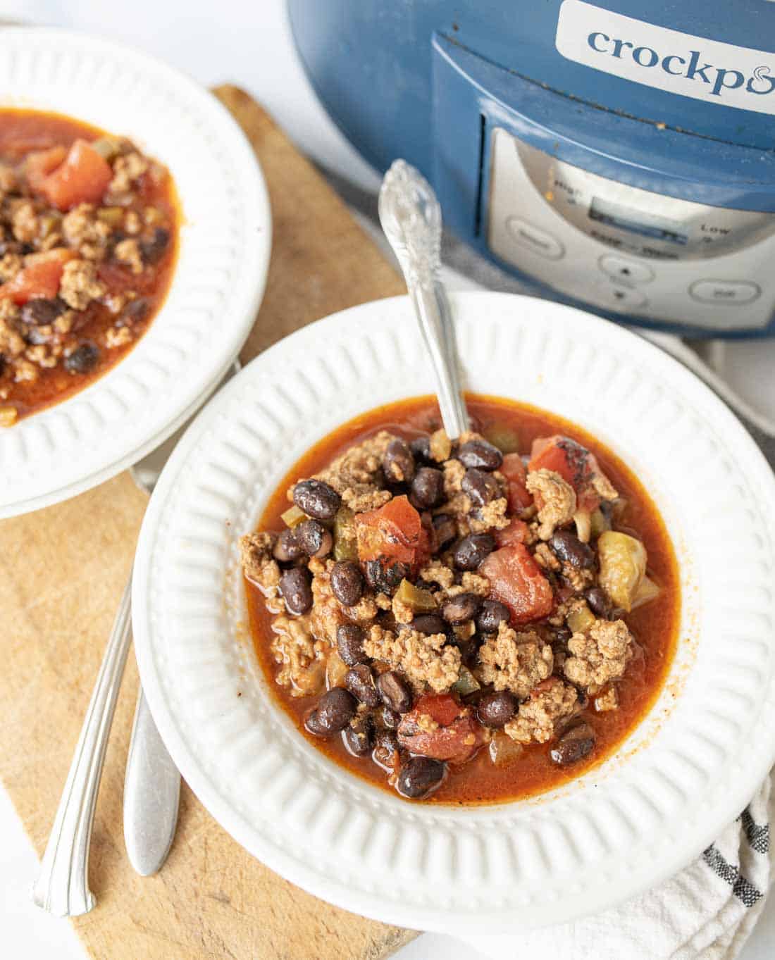 A bowl of black bean chili with ground meat, black beans, and tomatoes sits on a white plate next to a slow cooker labeled crockpot. A spoon is in the bowl and another bowl of chili is nearby.