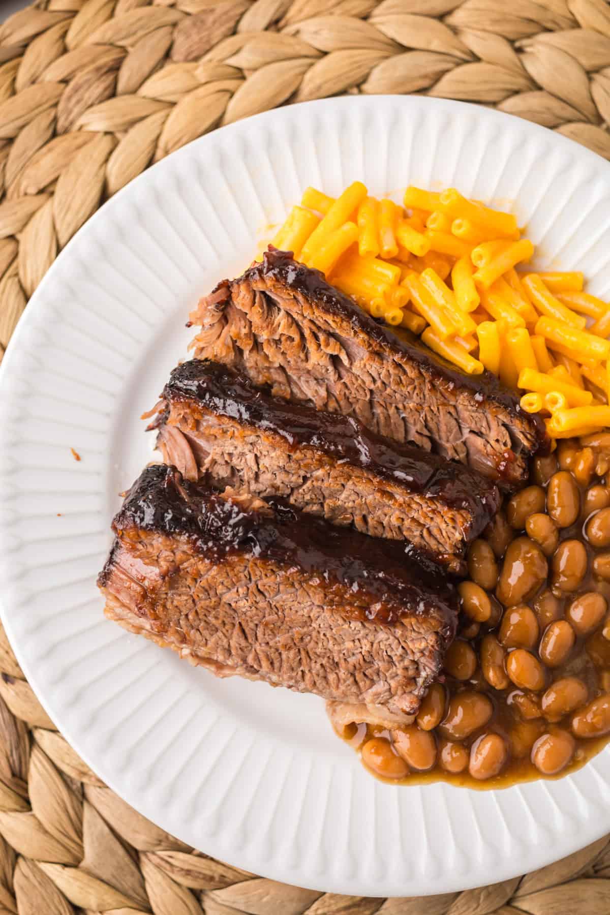 A white plate with slices of slow cooker brisket, baked beans, and yellow macaroni and cheese, placed on a woven placemat.