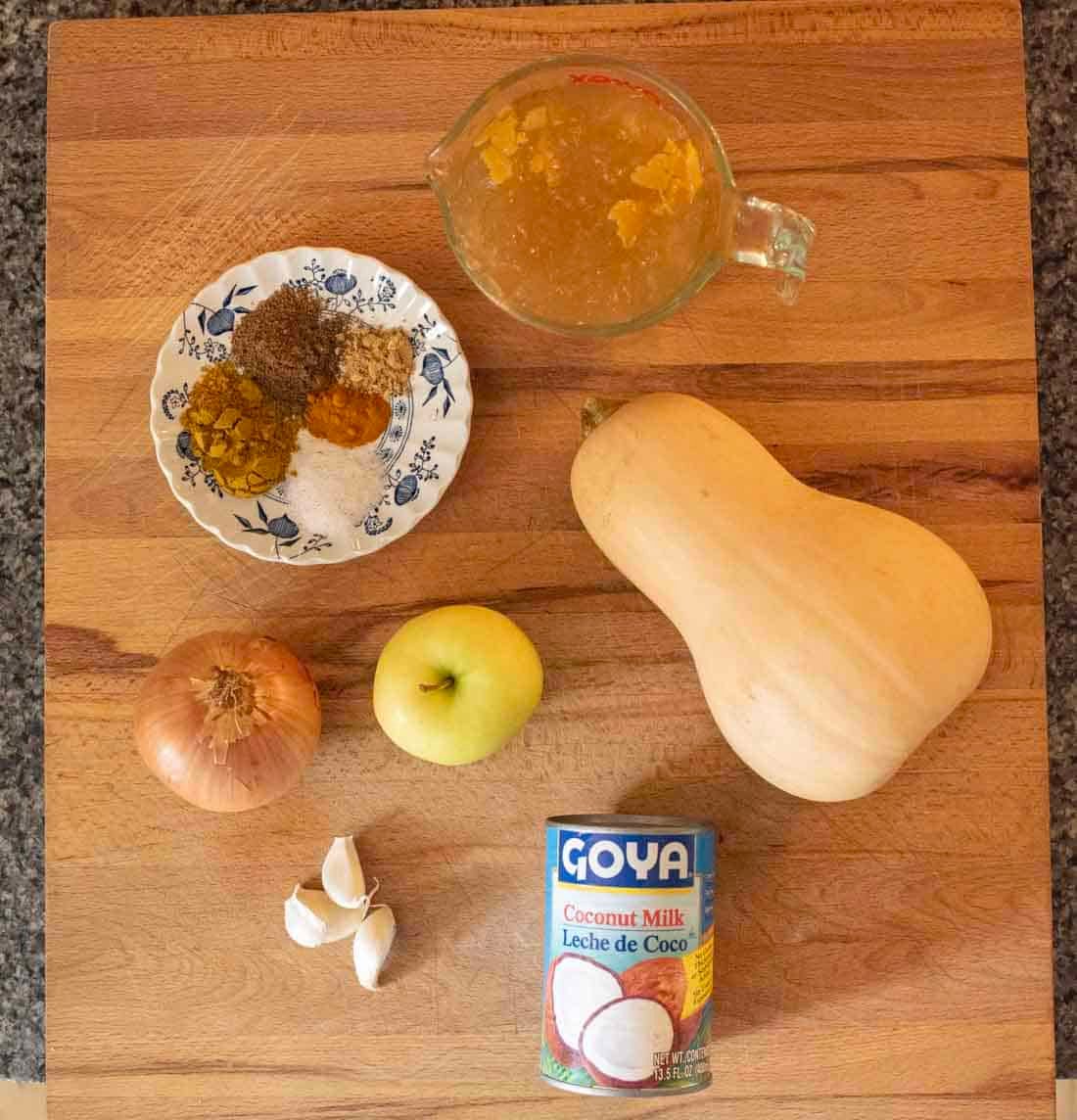 Ingredients for a recipe displayed on a wooden surface: a measuring cup with liquid, a plate with spices, a butternut squash, an onion, a green apple, garlic cloves, and a can of Goya coconut milk.