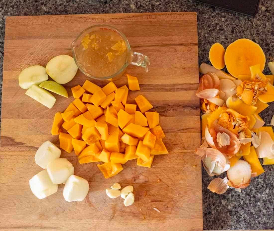 A wooden cutting board with chopped butternut squash, apple slices, peeled onions, garlic cloves, a measuring cup of broth, and a pile of vegetable peels on the side, set on a granite countertop.