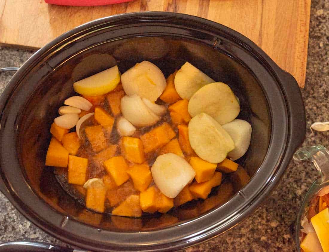 A slow cooker filled with cubed butternut squash, chunks of onion, sliced apples, and whole garlic cloves, ready to be cooked.