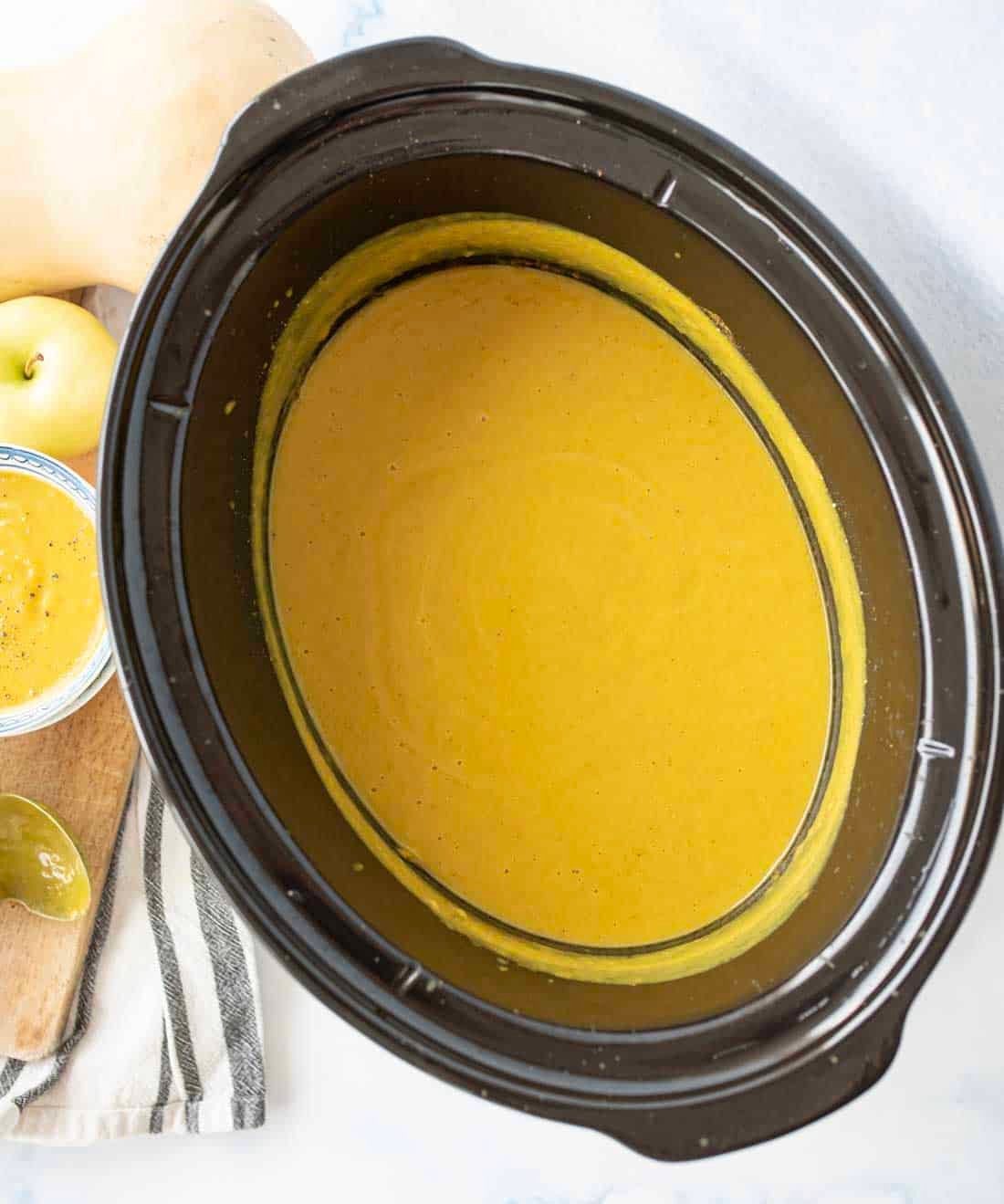 A slow cooker filled with smooth, creamy orange soup sits on a countertop next to a striped kitchen towel, a wooden board, a yellow apple, and part of a butternut squash.