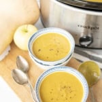 Two bowls of creamy slow cooker butternut squash soup, topped with black pepper, sit on a wooden board next to two spoons, a slow cooker, an apple, and a butternut squash. The slow cooker is filled with more soup.
