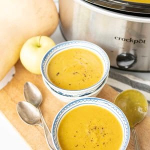 Two bowls of creamy slow cooker butternut squash soup, topped with black pepper, sit on a wooden board next to two spoons, a slow cooker, an apple, and a butternut squash. The slow cooker is filled with more soup.