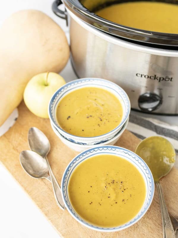 Two bowls of creamy slow cooker butternut squash soup, topped with black pepper, sit on a wooden board next to two spoons, a slow cooker, an apple, and a butternut squash. The slow cooker is filled with more soup.