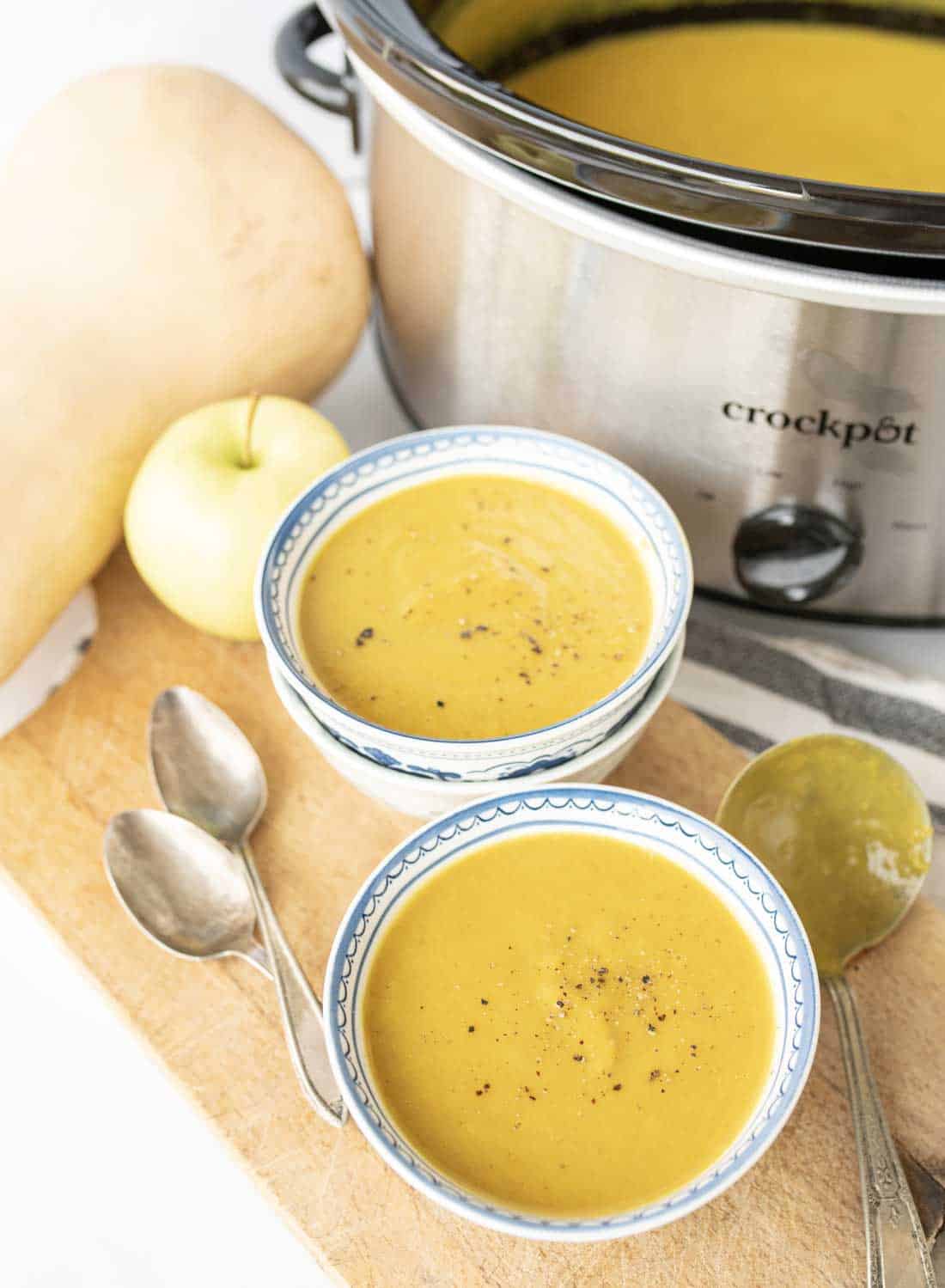 Two bowls of creamy slow cooker butternut squash soup, topped with black pepper, sit on a wooden board next to two spoons, a slow cooker, an apple, and a butternut squash. The slow cooker is filled with more soup.