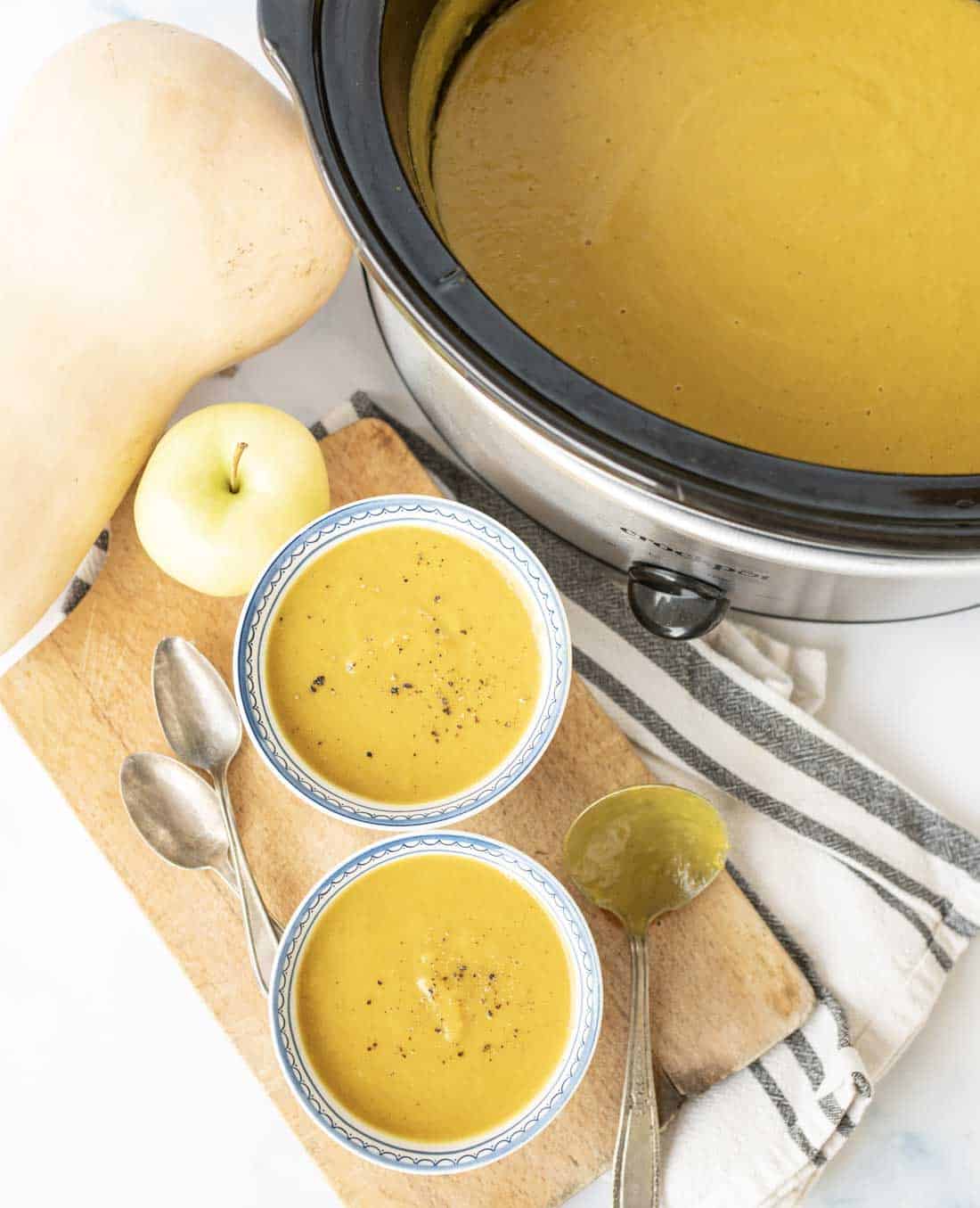 A slow cooker filled with creamy orange soup sits beside a cutting board with two bowls of soup, a butternut squash, an apple, two spoons, and a striped towel. Soup in bowls is topped with black pepper.