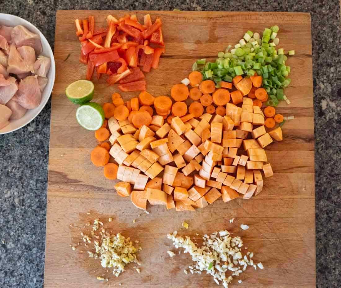 Chopped sweet potatoes, carrots, green onions, and red bell peppers are arranged on a wooden cutting board, with minced garlic and ginger in small piles and a halved lime. A plate of raw chicken sits to the side.