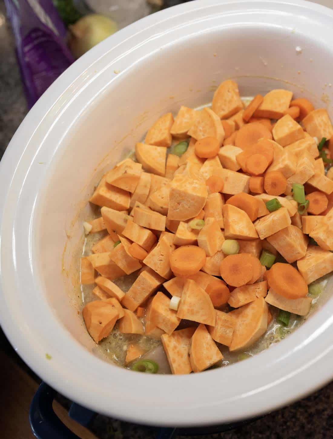 Chopped sweet potatoes, carrots, and green onions in a white slow cooker, ready to be cooked.