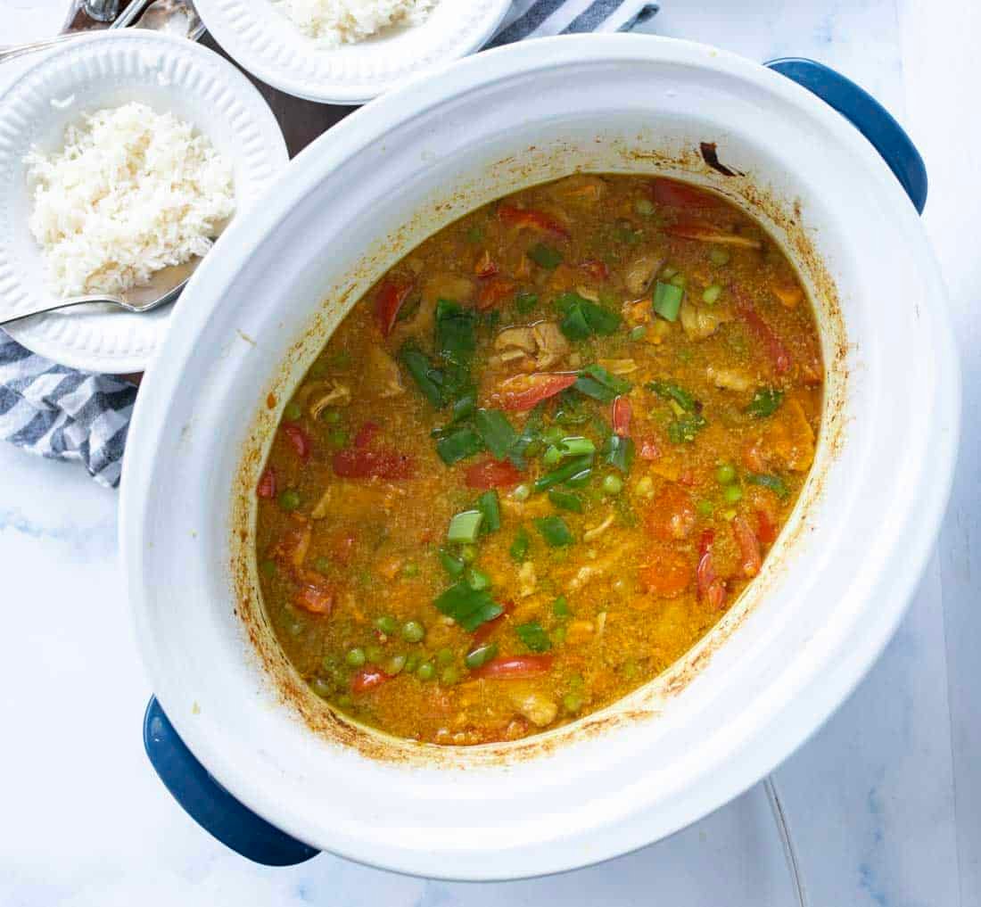 A white slow cooker filled with yellow chicken curry, garnished with green onions and surrounded by plates of white rice on a marble surface.