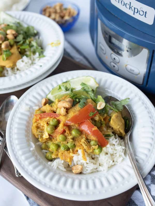 A plate of rice topped with chicken in yellow curry, vegetables, and peanuts, garnished with cilantro and lime, is served with a spoon. Another plate and a Crockpot slow cooker are visible in the background.