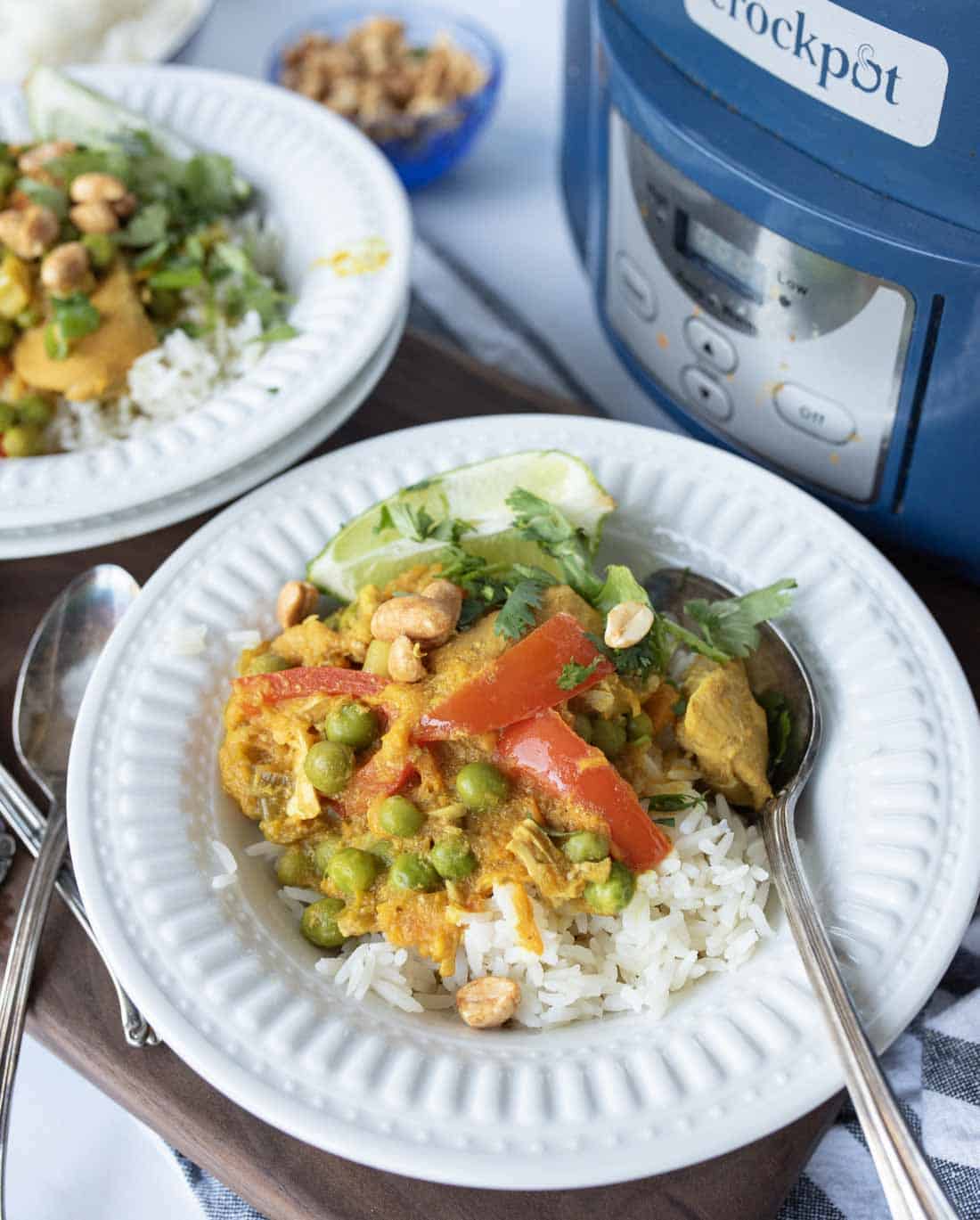 A plate of rice topped with chicken in yellow curry, vegetables, and peanuts, garnished with cilantro and lime, is served with a spoon. Another plate and a Crockpot slow cooker are visible in the background.